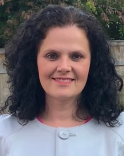 A white woman with curly black hair and a white shirt is smiling at the camera.