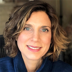 A white woman is leaning slightly to the side and smiling at the camera. She has chin-length wavy brown hair and blue eyes and is wearing a dark blue shirt