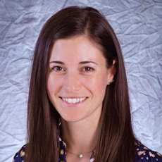 A young white woman is smiling at the camera. She has long straight dark hair and is wearing a floral shirt.