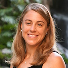 A woman with long wavy light brown hair is smiling at the camera. Her head is tilted to one side and she is wearing a black tank top.