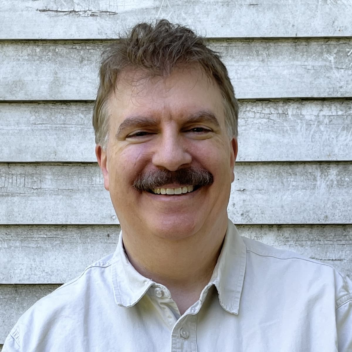A white man with short hair and a mustache, wearing a button up shirt, is looking at the camera and smiling.